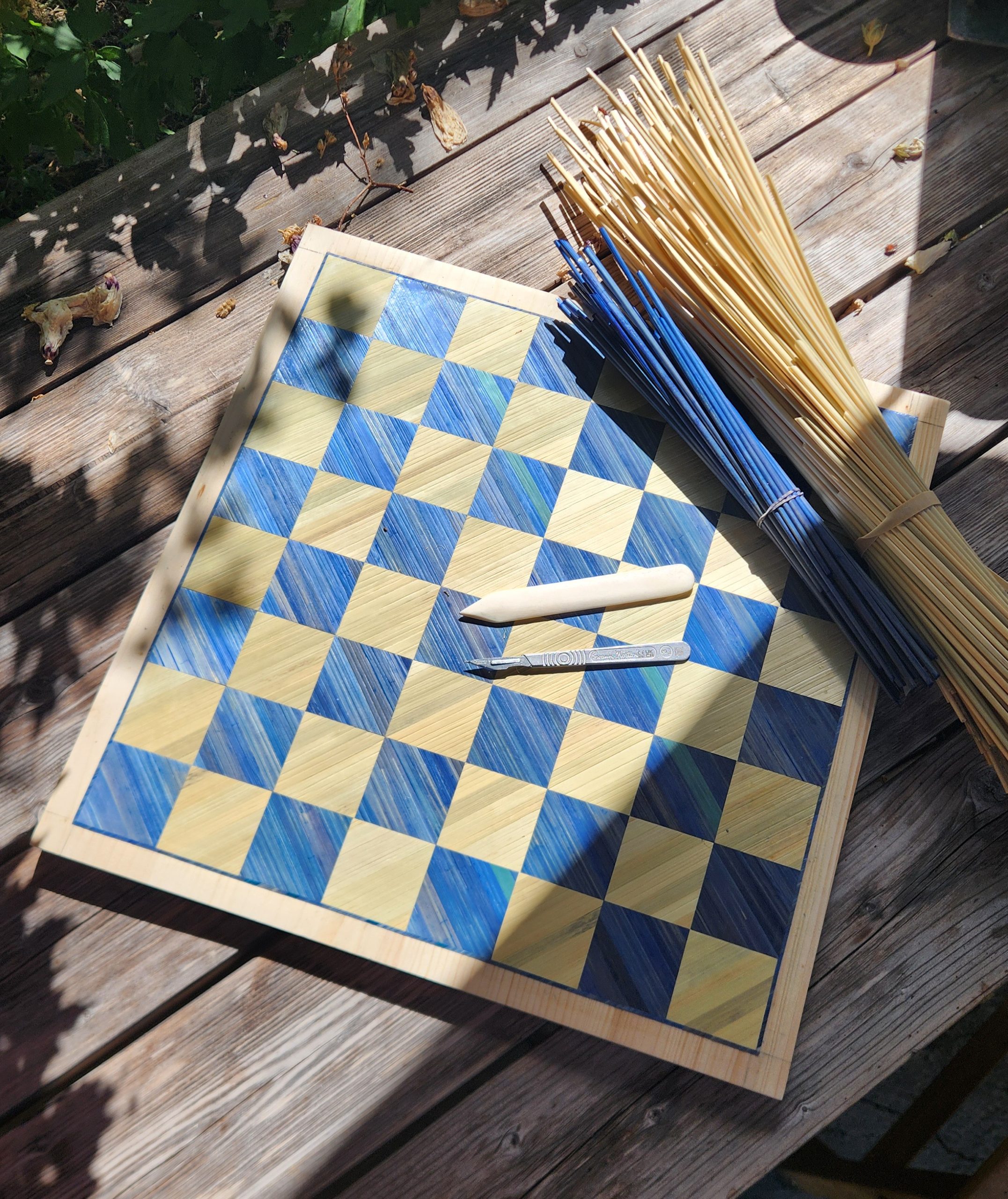 Échiquier en marqueterie de paille de seigle bleue et naturelle en cours de réalisation, avec plioir, scalpel et bottes de paille